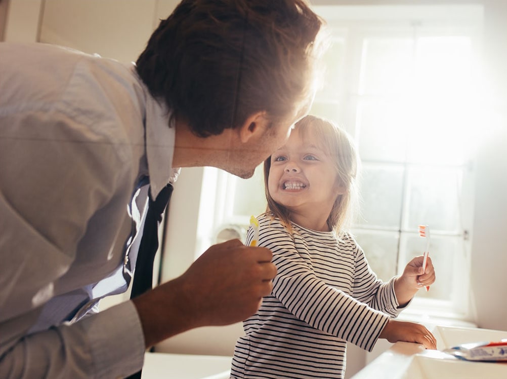 An adult and a young child stand by a sink holding toothbrushes. The child smiles widely at the adult, showing her teeth, with sunlight streaming through a window behind them.