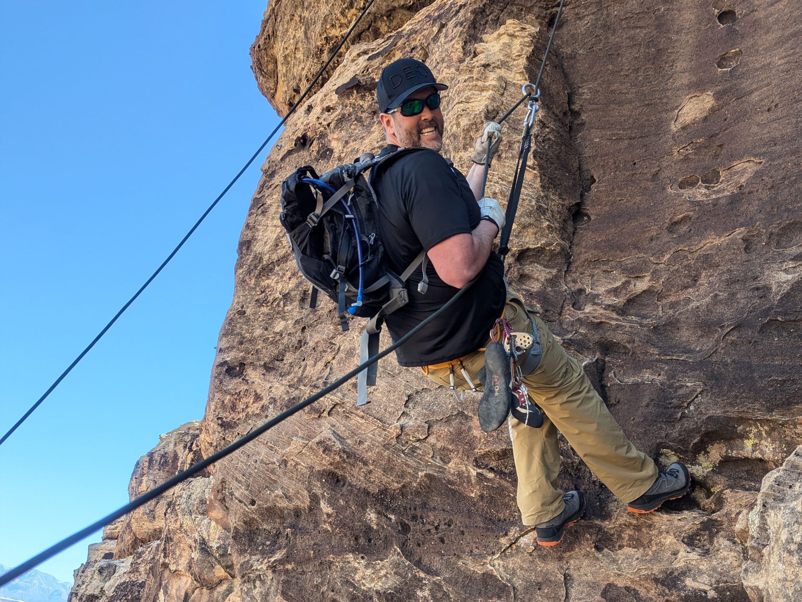 A man wearing climbing gear and sunglasses is rappelling down a steep rock face, secured by multiple ropes, with a blue sky in the background.
