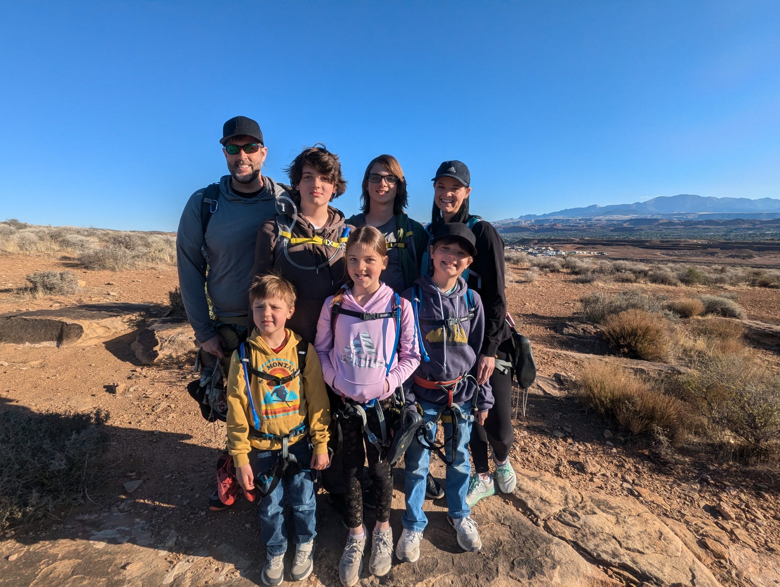 A group of seven people, including adults and children, dressed in outdoor gear, pose on a rocky desert landscape with mountains in the background under a clear blue sky.