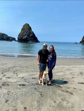 A man and woman stand on a sandy beach with a dog, with large rocks and the ocean in the background under a clear sky.