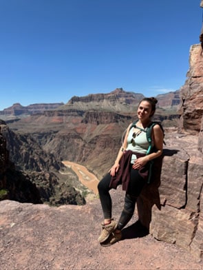 A woman stands on a rocky ledge overlooking the Grand Canyon with the Colorado River visible below under a clear blue sky.