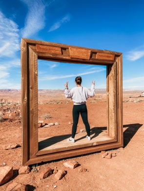 A person stands inside a large, wooden picture frame set in a desert landscape, facing away and making peace signs with both hands.