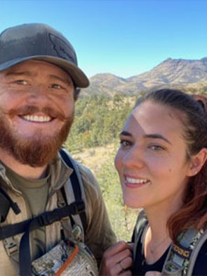 Two people with backpacks pose for a selfie outdoors with trees and mountains in the background on a sunny day.