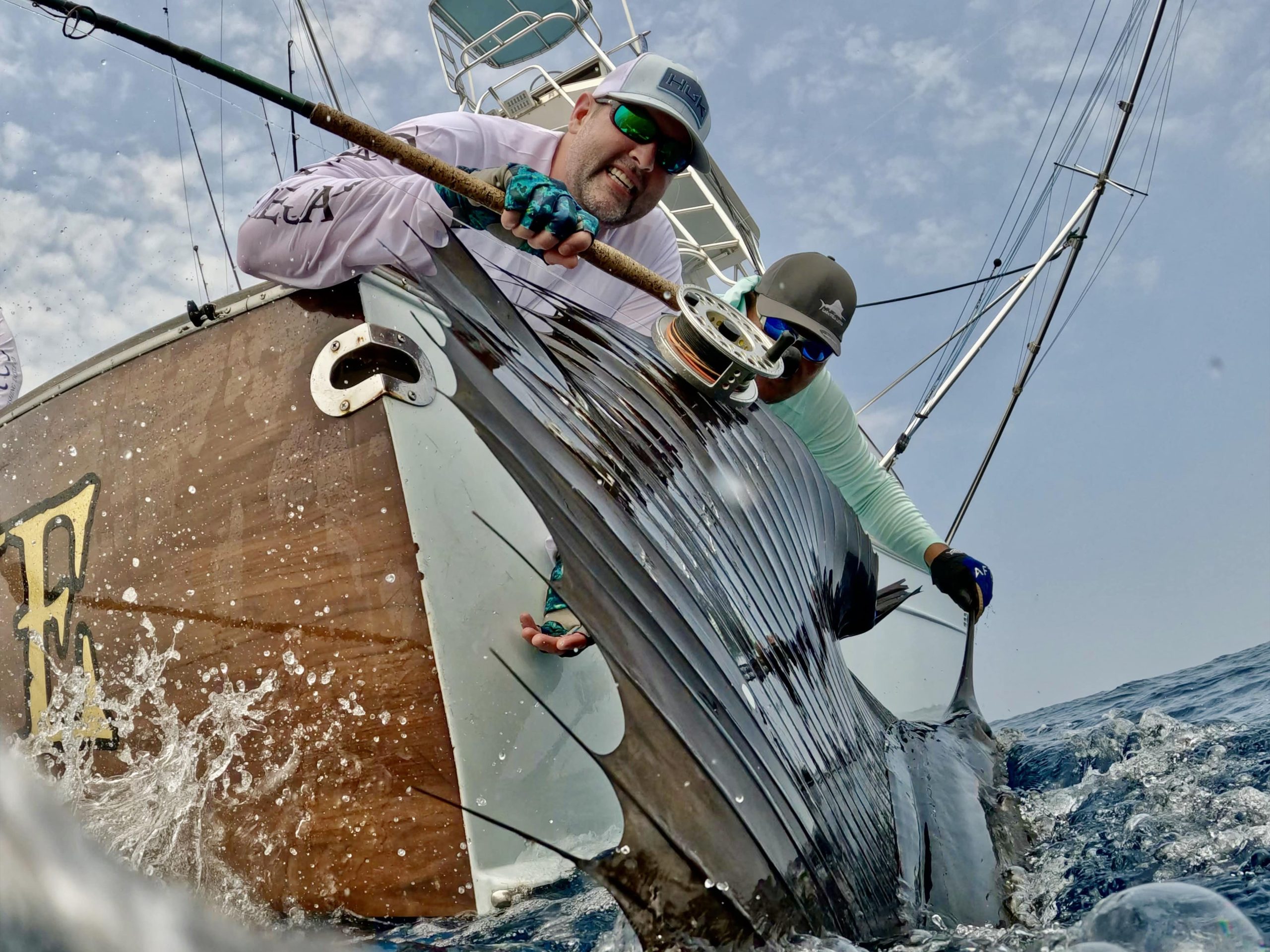 Two people on a boat hold a large sailfish partially out of the water by its bill, with fishing rods and ocean spray visible.