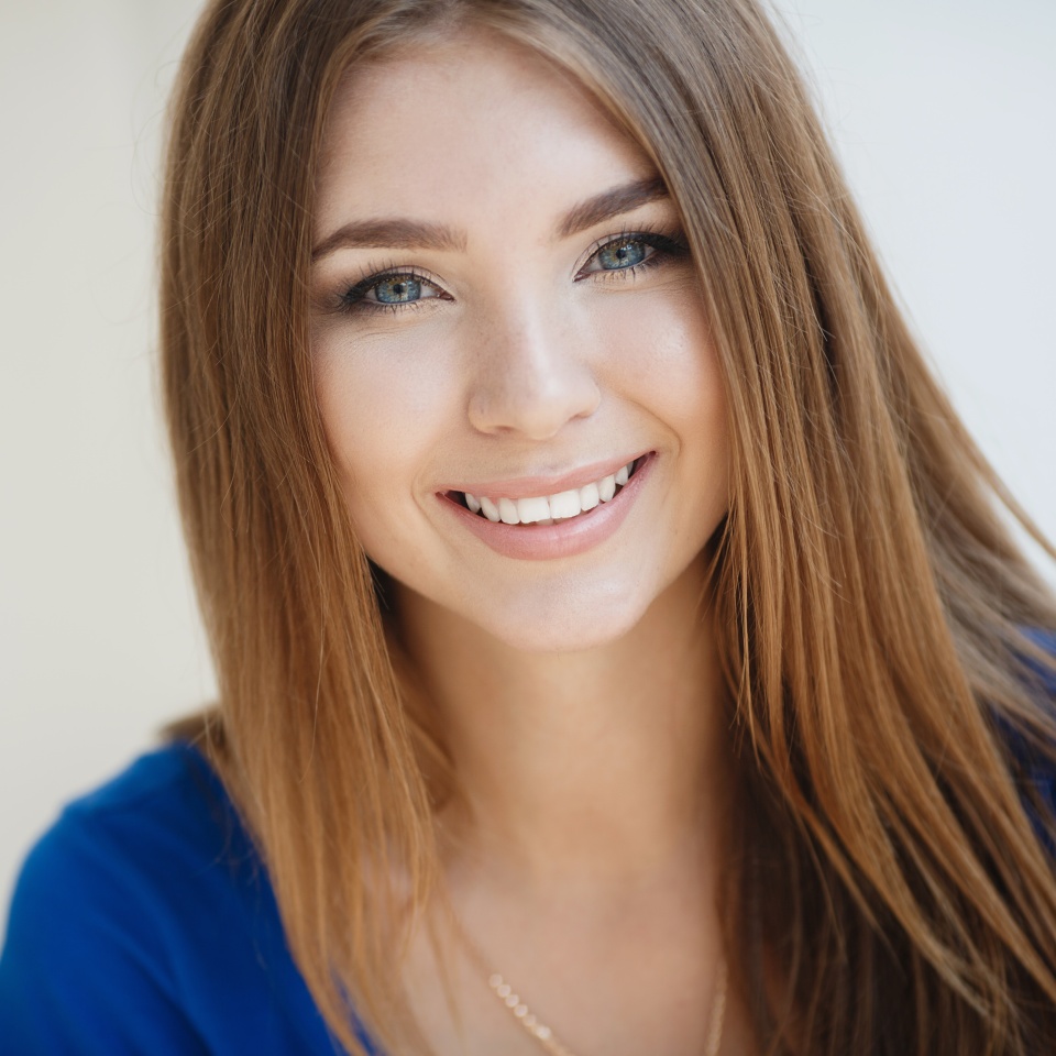 Beautiful young woman brunette with long straight hair,long black eyelashes,pretty smile,white straight teeth,wearing a blue summer dress,wears a gold chain on the neck,posing on light gray background