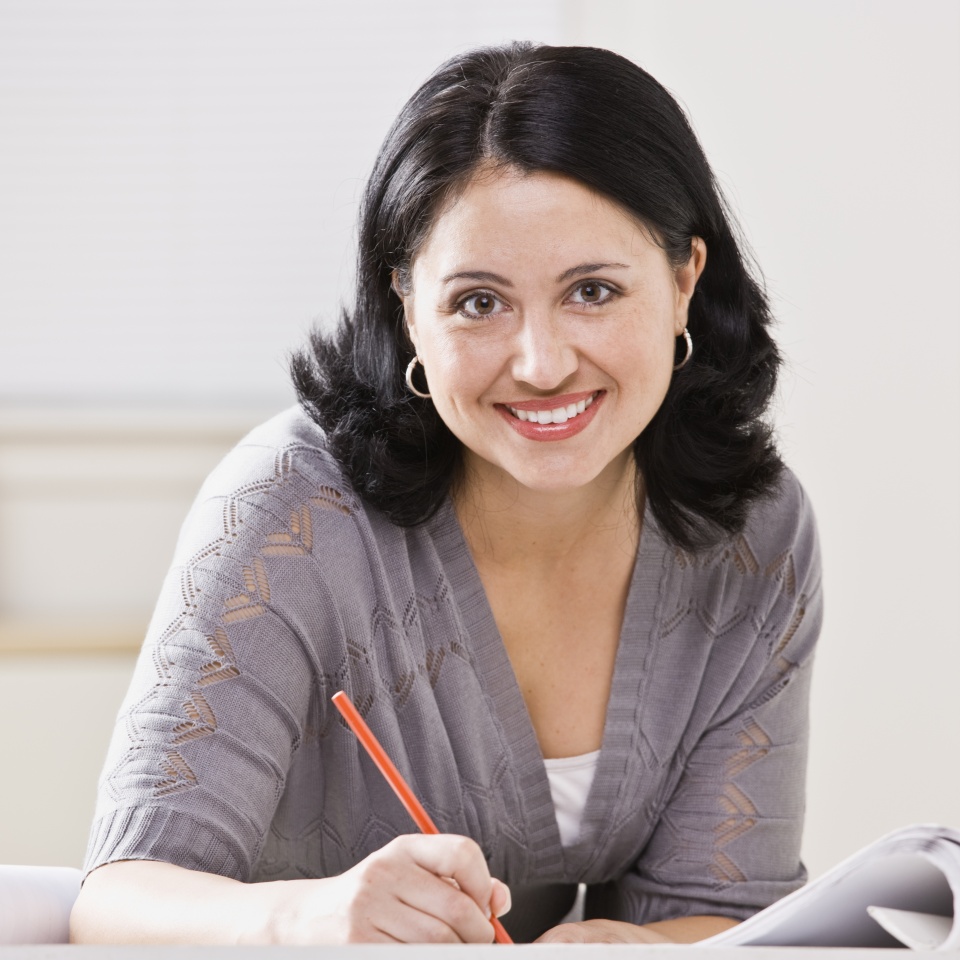 A beautiful Hispanic woman writing at a desk. She is smiling at the camera. Square compostion.