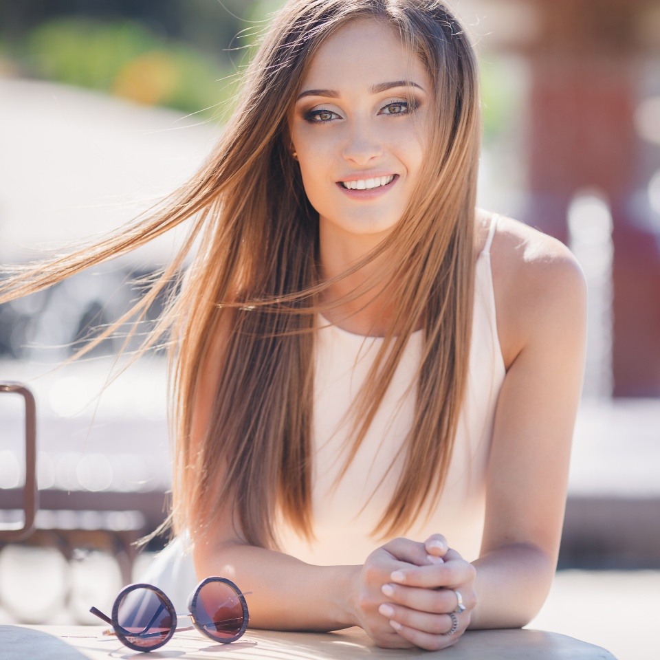 Young,beautiful,attractive woman,a brunette with long straight hair and gray eyes,a beautiful smile and straight white teeth,dressed in a white shirt,sitting at a table in a city cafe,posing against a green urban landscape.