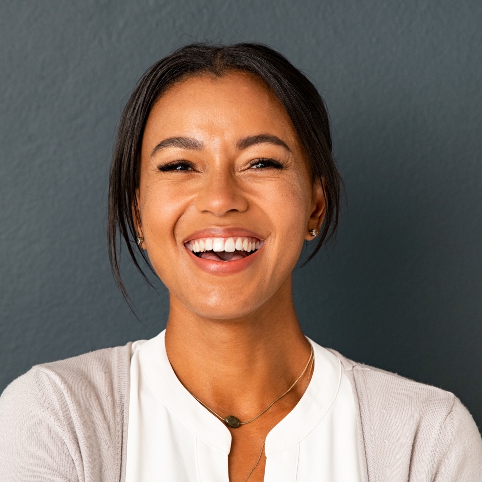 Close up face of beautiful mature woman looking at camera with big laugh. Mid adult hispanic woman standing isolated against grey wall while having fun. Portrait of smiling indian lady laughing.