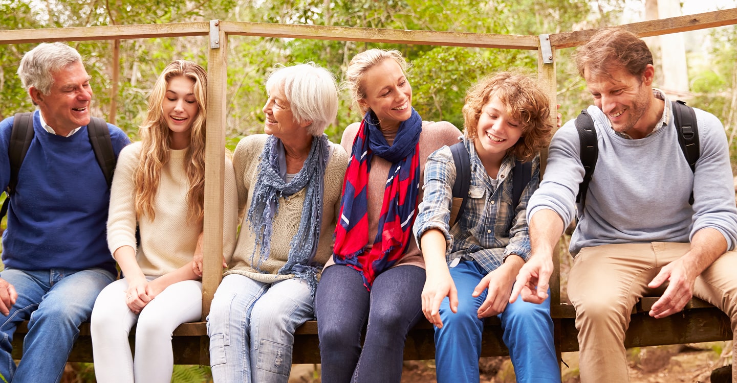 Six people of various ages sitting on a wooden bridge outdoors, smiling and interacting with each other, surrounded by greenery.