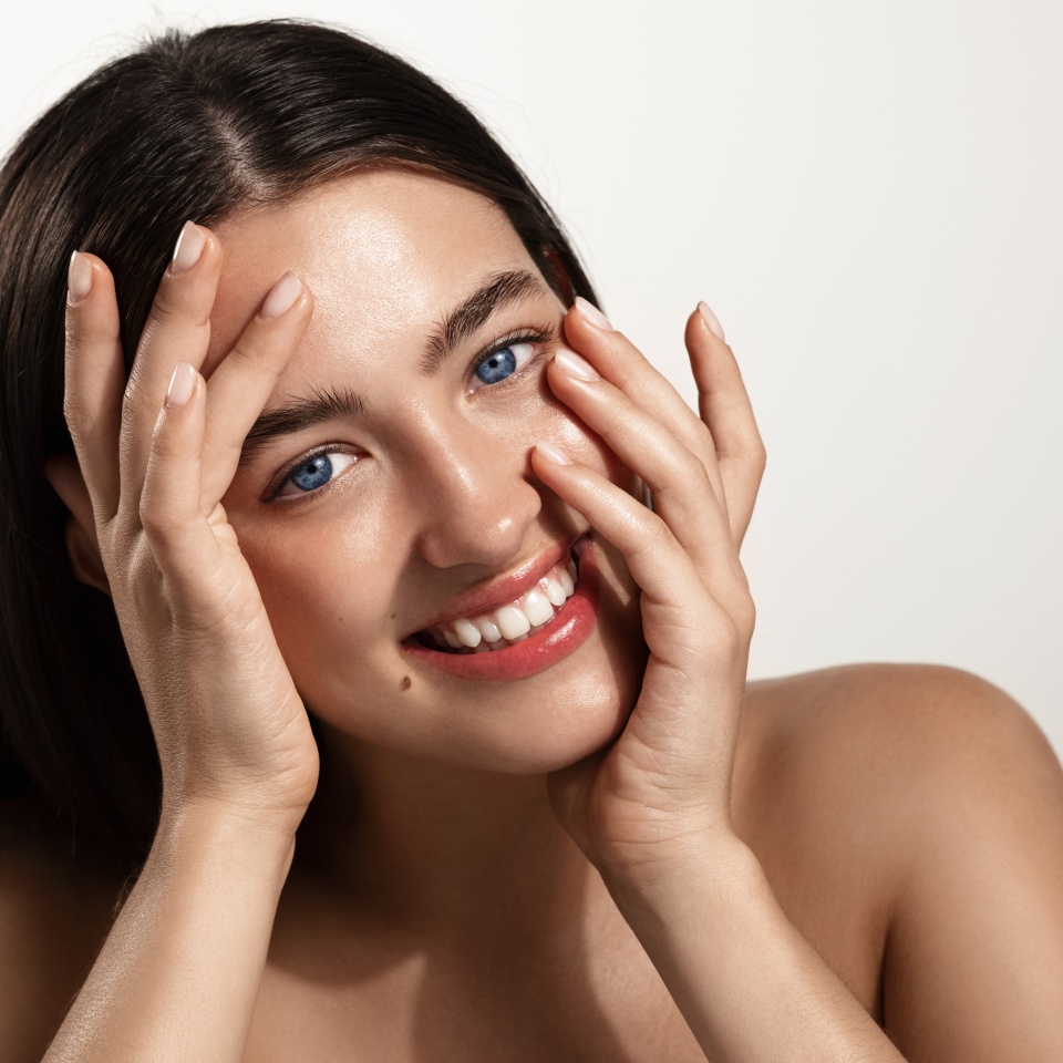 Radiant and glowing facial skin, women cosmetics. Beautiful girl model touching her nourished, hydrated clean face and smiling happy, blue eyes and healthy skin, white background