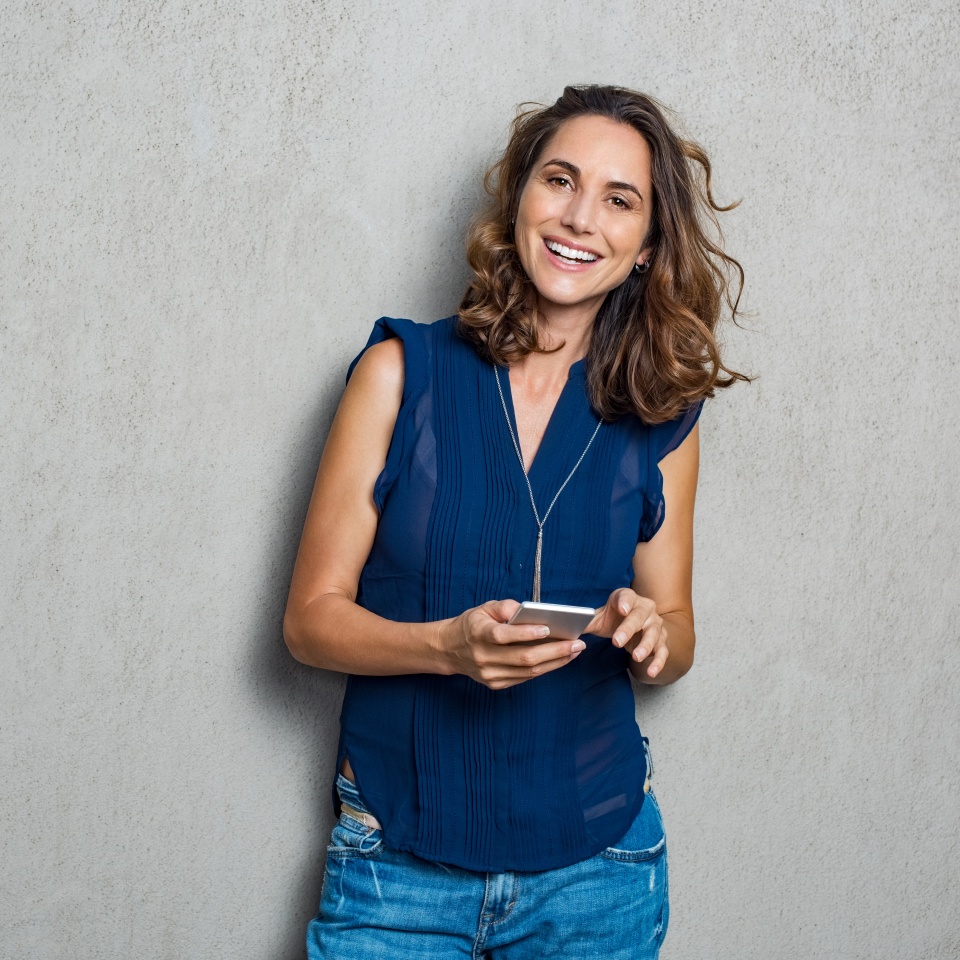 Smiling mature woman using smartphone and looking at camera. Happy woman typing on cellphone over gray background with copy space. Portrait of smiling latin woman messaging with smartphone.
