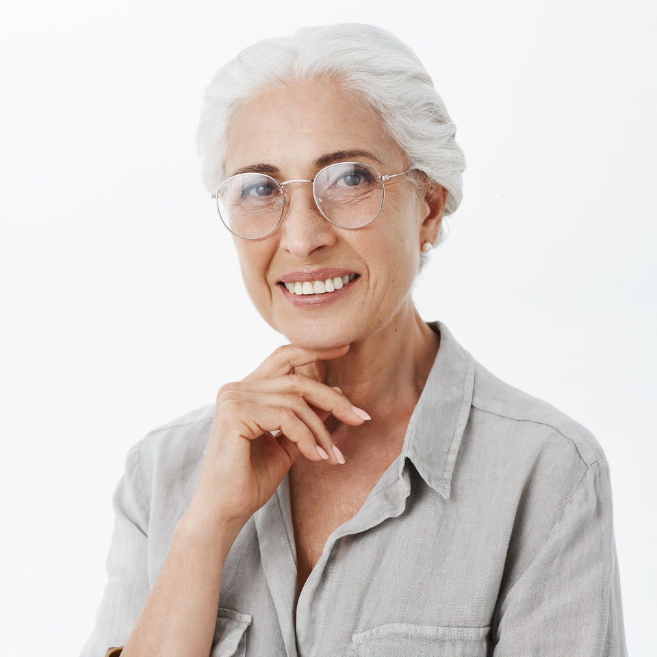 Waist-up shot of wise and pleased carefree kind grandmother in glasses with white hair holding hand on chin in thoughtful and delighted pose smiling broadly in satisfation and joy over gray wall