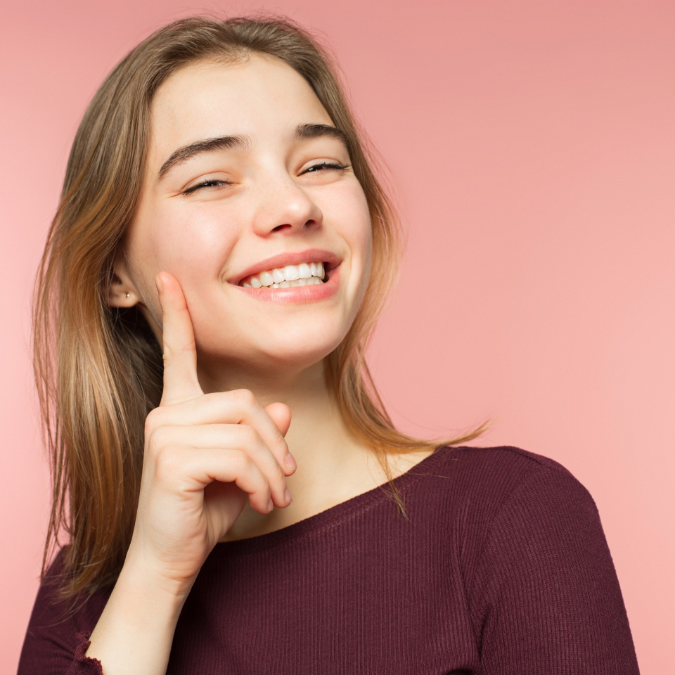Woman smiling with perfect smile and white teeth on the pink studio background and looking at camera. Pretty young girl. She smiles widely. Her hair is light brown. Happiness and youth concept. Front