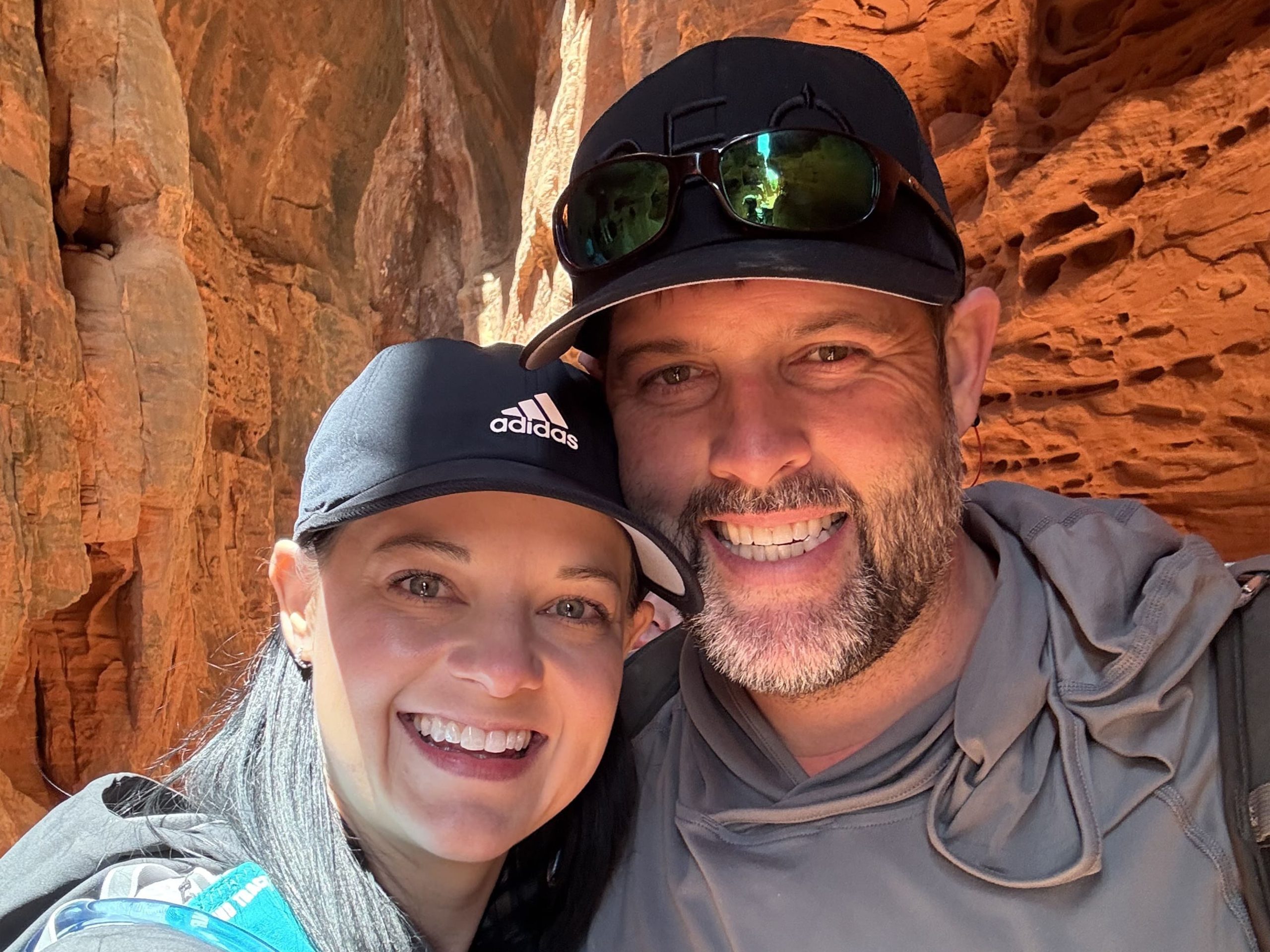 Two people wearing hiking attire smile for a close-up photo in front of red rock formations in a sunlit outdoor setting.