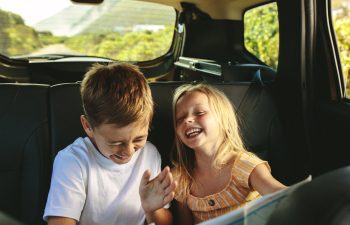 Two young children, a boy and a girl, sit in the backseat of a car, laughing and smiling together during a car ride.