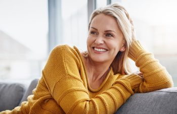A woman with blonde hair wearing a yellow sweater is sitting on a couch, smiling and looking to the side.