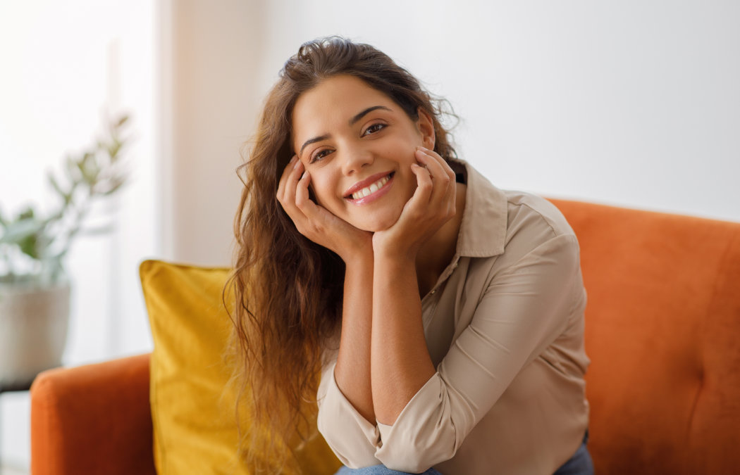 A woman with long brown hair smiles while sitting on an orange couch, leaning forward with her hands on her cheeks.