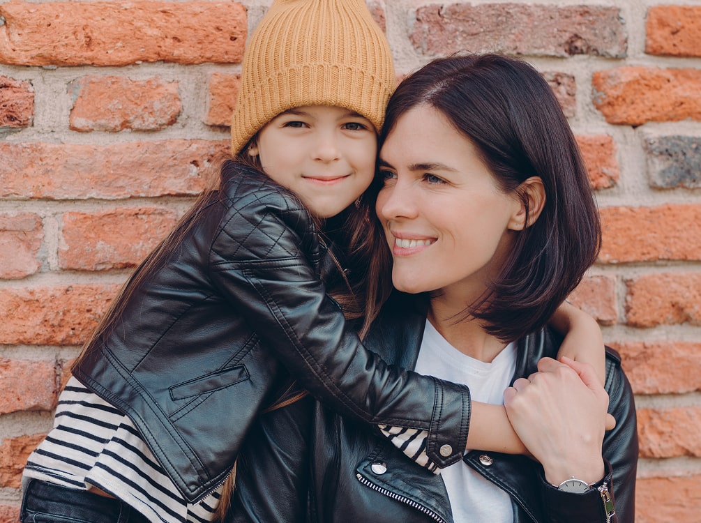A young girl wearing a yellow knit beanie hugs a woman in front of a brick wall; both wear black leather jackets and striped shirts.