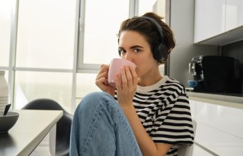Person sitting in a kitchen wearing headphones and drinking from a pink mug
